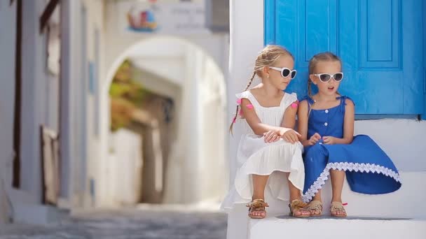 Twee meisjes in blauwe jurken plezier buitenshuis. Kinderen op straat van typische Griekse traditionele dorp met witte wanden en kleurrijke deuren op het eiland Mykonos, in Griekenland — Stockvideo