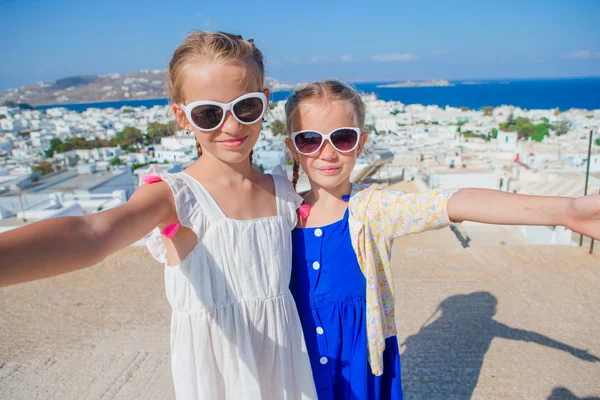 Dos chicas en vestidos azules tomando fotos de selfies al aire libre. Fondo para niños del típico pueblo tradicional griego con paredes blancas y puertas coloridas en la isla de Mykonos, en Grecia —  Fotos de Stock