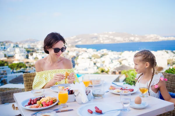 Familia desayunando en la cafetería al aire libre con una vista increíble de la ciudad de Mykonos. Adorable chica y mamá beber jugo fresco y comer croissant en la terraza del hotel de lujo —  Fotos de Stock