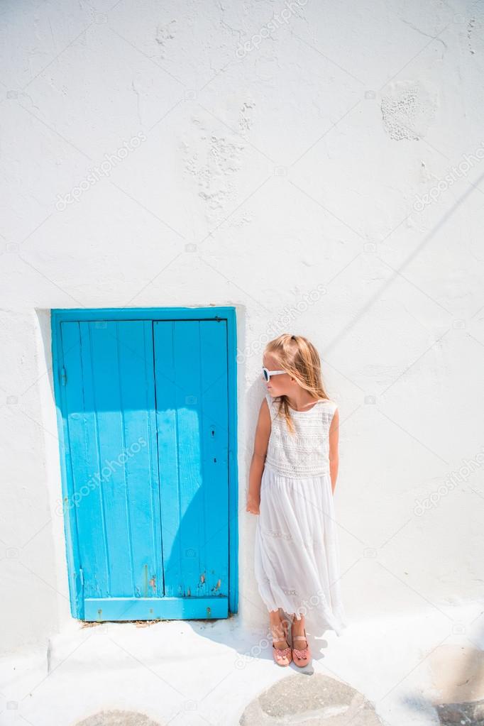 Charming girl in white dress outdoors in old streets an Mykonos. Kid at street of typical greek traditional village with white walls and colorful doors on Mykonos Island, in Greece