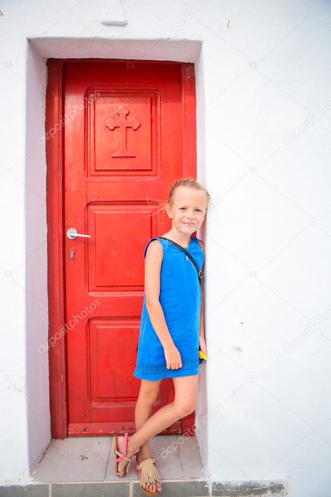 Cute girl in blue dress having fun outdoors near church. Kid at street of typical greek traditional village with white walls and colorful doors on Mykonos Island, in Greece