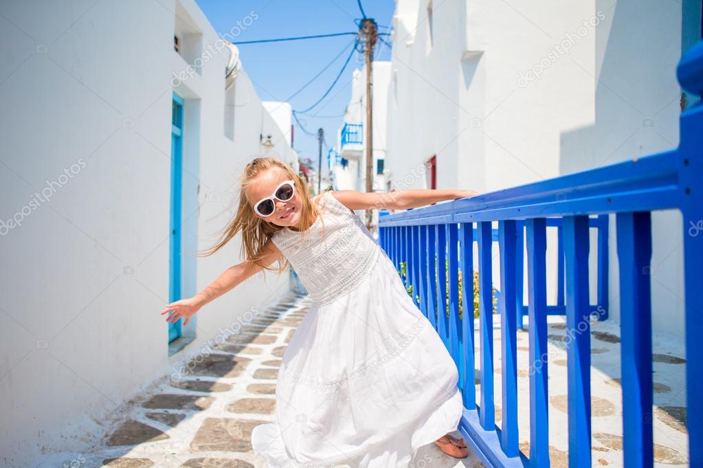 Charming girl in white dress outdoors in old streets an Mykonos. Kid at street of typical greek traditional village with white walls and colorful doors on Mykonos Island, in Greece