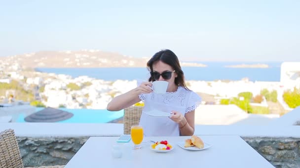 Linda senhora elegante tomando café da manhã no café ao ar livre com vista incrível sobre a cidade de Mykonos. Mulher bebendo café quente no terraço do hotel de luxo com vista para o mar no restaurante resort . — Vídeo de Stock