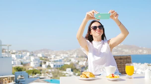 女性モバイルのスマート フォンを使用した朝食の写真を撮影。ソーシャル メディアのための贅沢な旅行休暇に食べ物の写真を撮る女の子。ミコノス島、ギリシャ、ヨーロッパのリゾート地に美しい女性. — ストック動画