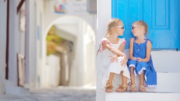 Δύο κορίτσια στο μπλε φορέματα που διασκεδάζουν σε εξωτερικούς χώρους. Παιδιά στο δρόμο της τυπικό ελληνικό παραδοσιακό χωριό με λευκούς τοίχους και πολύχρωμες πόρτες στην Μύκονο, στην Ελλάδα — Αρχείο Βίντεο