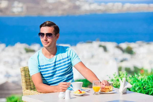 Young boy having breakfast at outdoor cafe with amazing view on Mykonos town. Man drinking hot coffee on luxury hotel terrace with sea view at resort restaurant. — Stock Photo, Image