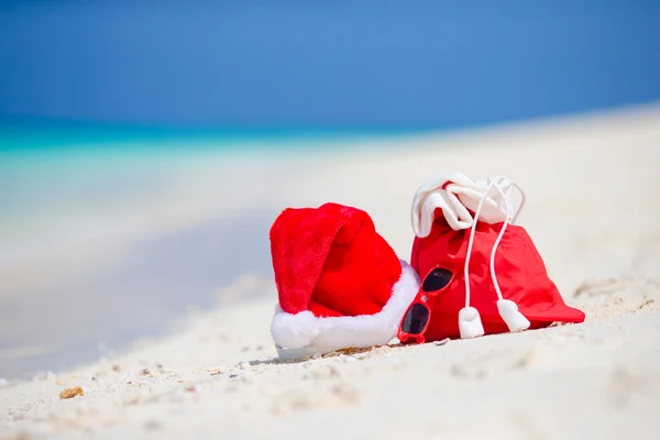 Closeup of red Santa bag and Santa Claus hat on beach. Xmas travel vacation and travel cuprise concept. Beach accessories with Santa Hat on white tropical beach — Stock Photo, Image