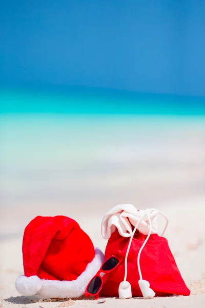 Zbliżenie czerwonej torby Mikołaja i kapelusz Świętego Mikołaja na plaży. Świąteczne wakacje podróży i podróży Cuprise koncepcji. Akcesoria plażowe z Santa Hat na białej tropikalnej plaży — Zdjęcie stockowe