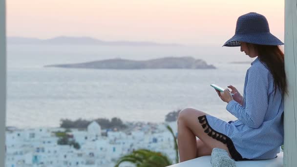 Mujer joven enviando un mensaje al atardecer sobre un mar en el famoso pueblo de Mykonos con los edificios antiguos en el fondo. Luz de fondo suave verano . — Vídeo de stock