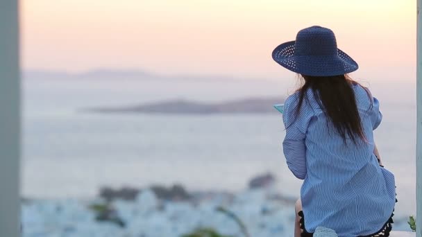 若い女性が背景に古い建物と有名な村ミコノス島の海の上の夕日を見ています。柔らかい夏のバックライト. — ストック動画