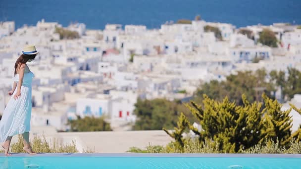 Hermosa joven que se relaja cerca de la piscina con una vista increíble de Mykonos. Chica de pie en el borde de la piscina infinita en Grecia — Vídeos de Stock