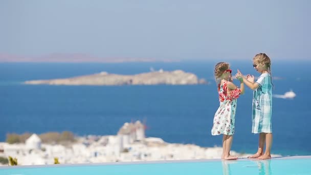 Schattige kleine meisjes spelen in de rand van het buitenzwembad met een prachtig uitzicht op de oude stad van Mykonos, Europa — Stockvideo