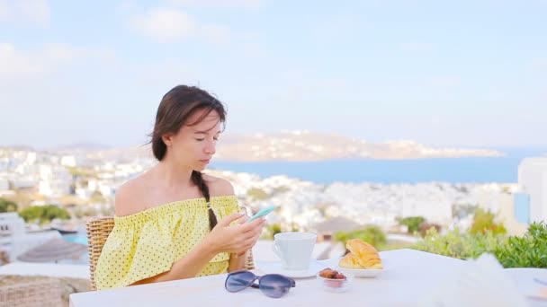 高加索女孩早餐与智能手机在户外咖啡馆与米科诺斯镇惊人的景色. — 图库视频影像