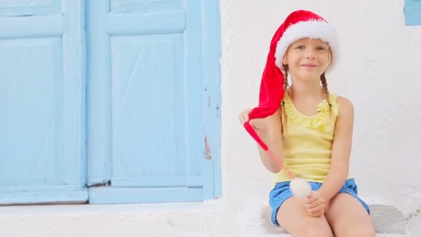 Маленькая девочка в шляпе Санты на улице на рождественских каникулах в Миконосе. Ребенок на улице типичной греческой традиционной деревни с белыми стенами и красочные двери на острове Миконос, в Греции — стоковое видео