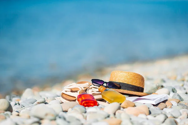 Пляжная шляпа на открытой книге с солнцезащитным кремом и обувью на галечном пляже — стоковое фото