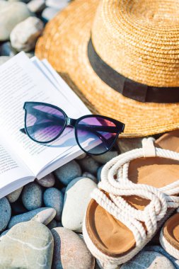 Plaj şapkası açık kitap, güneş kremi ve çakıl taşı plajında ayakkabılar.
