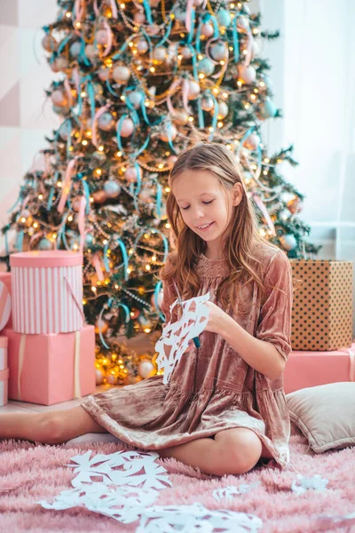 Чарівна маленька дівчинка сидить біля дерева і робить паперові сніжинки. Кімната прикрашена . — стокове фото