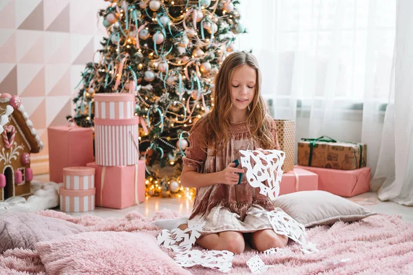 Entzückendes kleines Mädchen, das neben dem Baum sitzt und Papierschneeflocken bastelt. Raum dekoriert. — Stockfoto