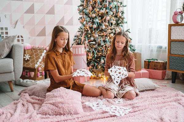 Entzückende kleine Mädchen, die neben dem Baum sitzen und Schneeflocken aus Papier herstellen. Raum dekoriert. — Stockfoto