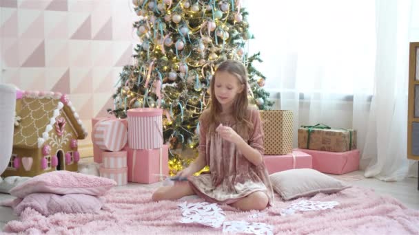 Urocza dziewczynka siedząca obok drzewa i robiąca papierowe płatki śniegu. Pokój urządzony. — Wideo stockowe