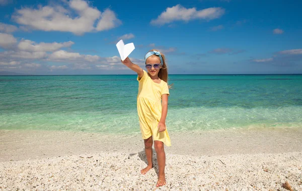 Adorable little girl with paper airplane in hands on white sandy beach — Stock Photo, Image