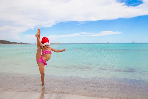 Smiling little girl making stretching exercise in Santa Hat on white beach — Stock Photo, Image