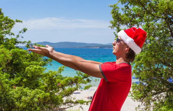 Giovane a Santa Hat sulla spiaggia tropicale vicino agli abeti — Foto Stock