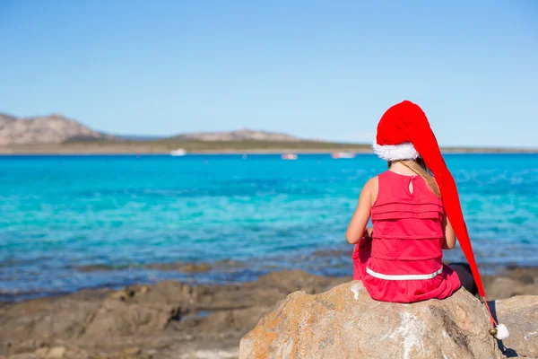 Adorabile bambina in cappello di Babbo Natale e vestito rosso seduto su grande pietra — Foto Stock