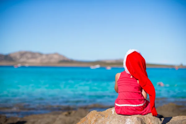 Adorable little girl in Santa hat and red dress sitting on big stone — Stock Photo, Image