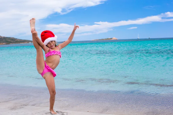 Lächelndes kleines Mädchen macht Dehnübungen in Weihnachtsmütze am weißen Strand — Stockfoto