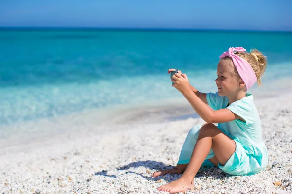 Little cute girl taking pictures on the phone at tropical beach — Stock Photo, Image