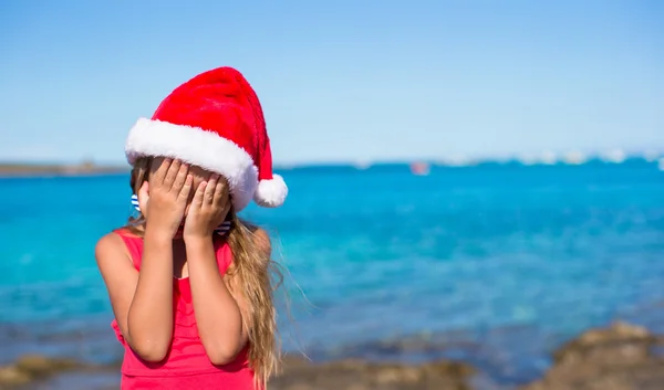 Little girl in santa hat covering her face background the sea — Stock Photo, Image