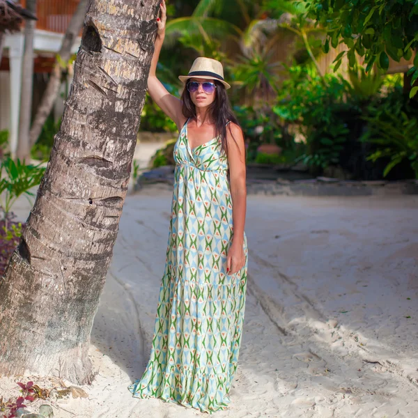 Beautiful brunette woman in long dress and hat relaxing near palmtree — Stock Photo, Image