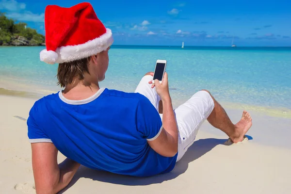 Widok młodego człowieka w santa hat z telefonu z powrotem na tropikalnej plaży — Zdjęcie stockowe