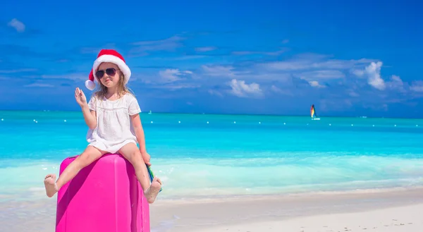 Kleines süßes Mädchen mit Weihnachtsmütze auf Koffer am tropischen Strand — Stockfoto