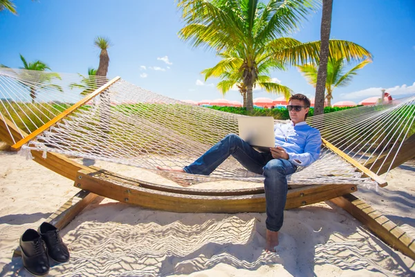 Mladý muž pracující s přenosným počítačem v houpací síti během pláž dovolená — Stock fotografie