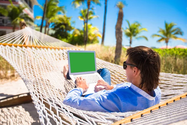 Młody mężczyzna pracujący z laptopa w hamaku podczas wakacji na plaży — Zdjęcie stockowe