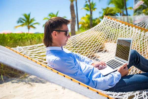 Молодий чоловік працює з ноутбуком в гамаку під час пляжних канікул — стокове фото
