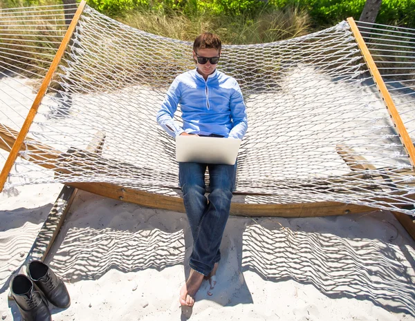 Młody mężczyzna pracujący z laptopa w hamaku podczas wakacji na plaży — Zdjęcie stockowe