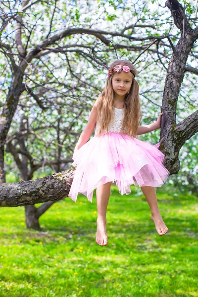 Liten bedårande flicka sitter på blommande träd i äppelträdgård — Stockfoto