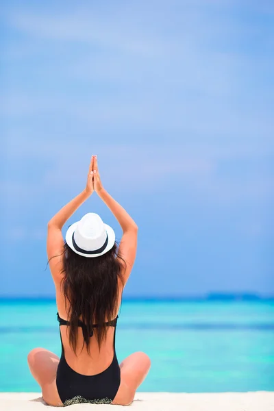 Healthy young woman in hat sitting in yoga position meditating on white sandy beach — Stock Photo, Image