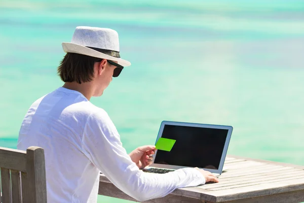 Young man working on laptop with credit card at tropical beach — Stock Photo, Image