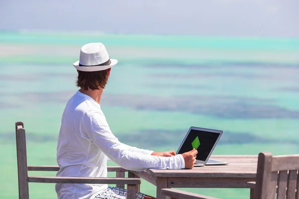 Young man working on laptop with credit card at tropical beach — Stock Photo, Image