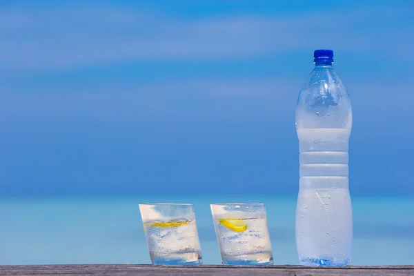 Glazen water met citroen en fles op houten op zee achtergrond — Stockfoto
