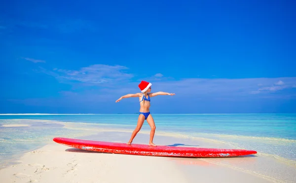 Adorable little girl in Santa hat practice surfing position on surfboard during vacation — Stock Photo, Image