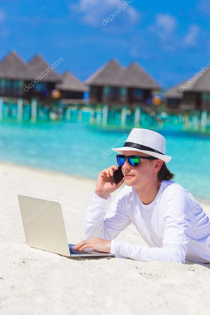Young business man with computer and cell phone on white beach