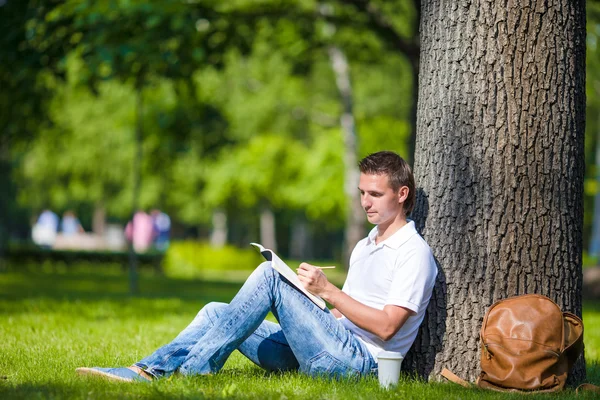 Jovem relaxante perto da árvore com café lendo um livro — Fotografia de Stock