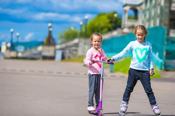 Two little girls roller skating and riding a scooter in the park outdoor — Stock Photo, Image