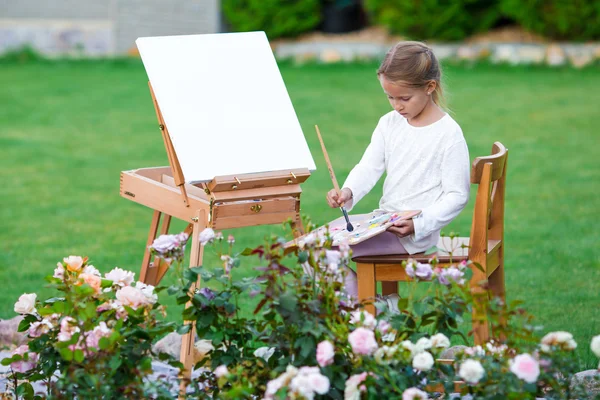 Adorável menina pintando um quadro em um cavalete em um dia quente ao ar livre — Fotografia de Stock