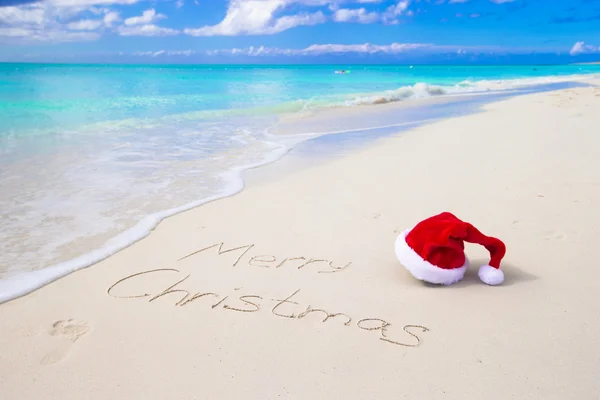 Frohe Weihnachten am Strand weißen Sand mit roter Weihnachtsmütze geschrieben — Stockfoto